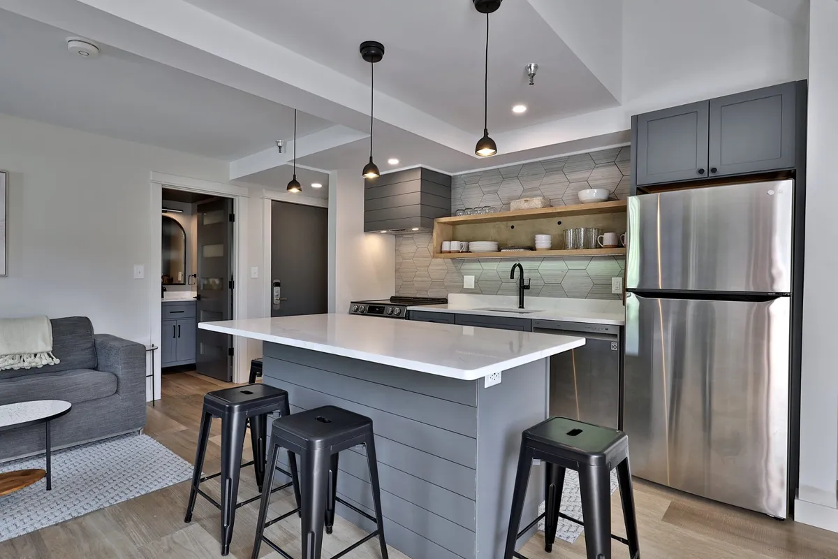 This image features a stylish, contemporary kitchen with a prominent island, stainless steel appliances, a hexagonal tile backsplash, and open wooden shelving. The island is spacious and inviting, ideal for meal prep and casual dining, while the overall design emphasizes a welcoming environment for cooking and socializing.