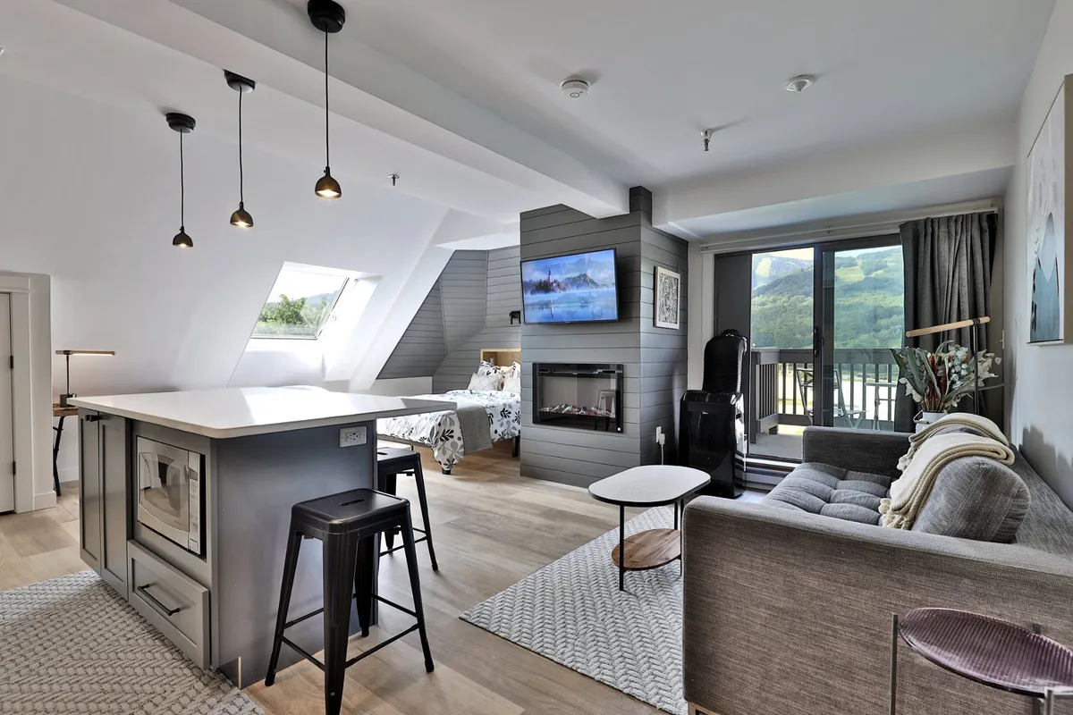 The hero image showcases a stylish, newly renovated studio condo interior featuring a modern kitchen island, inviting sofa by a cozy fireplace, and expansive windows framing breathtaking mountain views. The space is bathed in natural light, creating a warm and welcoming atmosphere ideal for relaxation after a day of outdoor adventures.