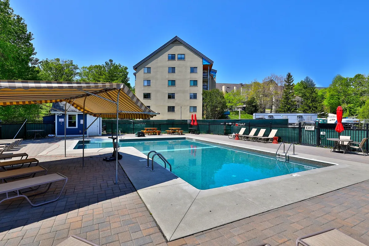 A sparkling outdoor swimming pool surrounded by lounge chairs and lush greenery. An umbrella shelters a few chairs, suggesting comfort for sunbathing, social gatherings, or reading in the shade. The setting is bright and leisurely, showcasing summer fun at the resort.