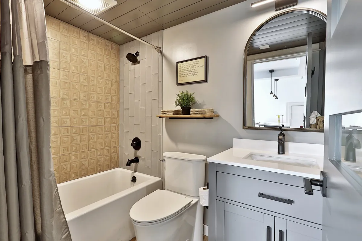 A serene modern bathroom with a bathtub, large mirror, vanity, subtle wall art, and a neatly arranged shelf with a potted plant. The ambient lighting and clean lines create an inviting space for relaxation and self-care rituals.