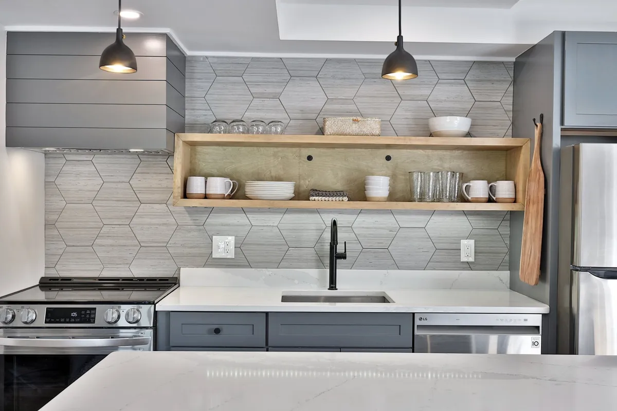 A close-up of the kitchen's hexagonal tile backsplash, open wooden shelves, sleek countertops, stainless steel appliances, and pendant lighting. The setup is warm and inviting, emphasizing both style and functionality for meal preparation and gatherings.