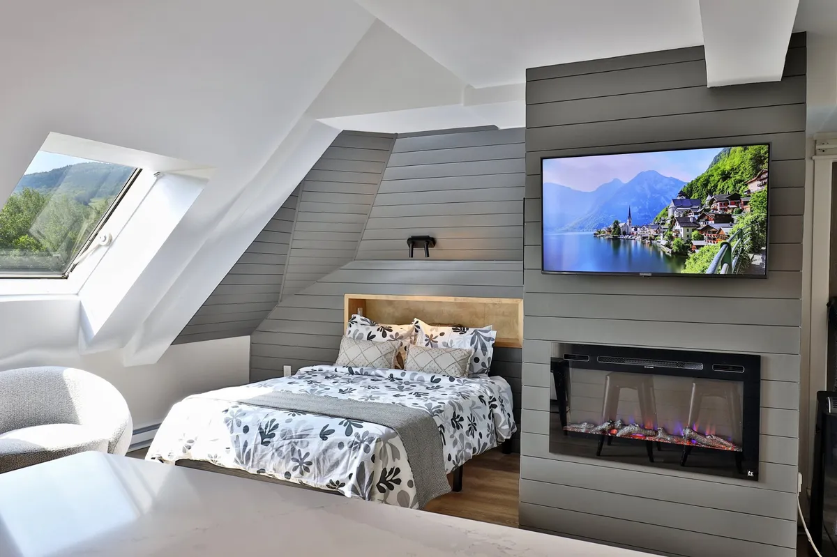 A serene corner of the studio with a queen bed dressed in floral bedding, a mounted smart TV, an electric fireplace, and a large window with views. The arrangement is cozy, inviting relaxation and entertainment with scenic surroundings.