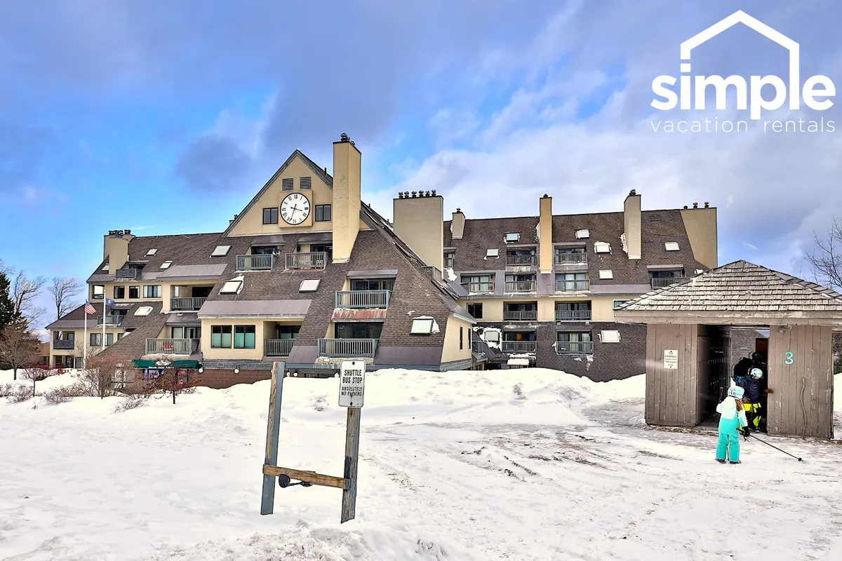This exterior image showcases the entrance to the building at Mountain Green, with its charming clock tower, snow-covered ground, and people dressed in winter attire, suggesting active engagement with the winter environment right outside the property.