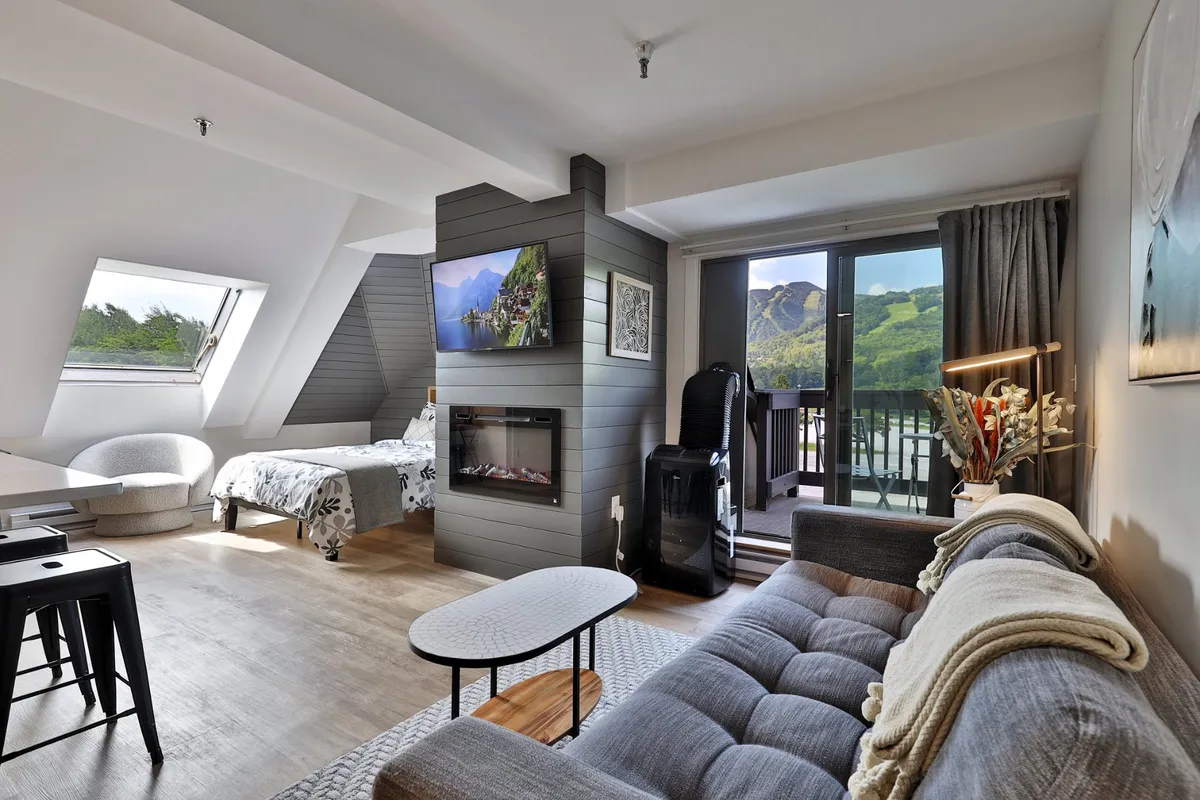 A spacious open-concept studio living area with a plush queen bed, modern sofa, and an electric fireplace beneath a wall-mounted smart TV. Floor-to-ceiling windows provide mountain views and access to a private balcony, filling the room with natural light and inviting the outdoors in.