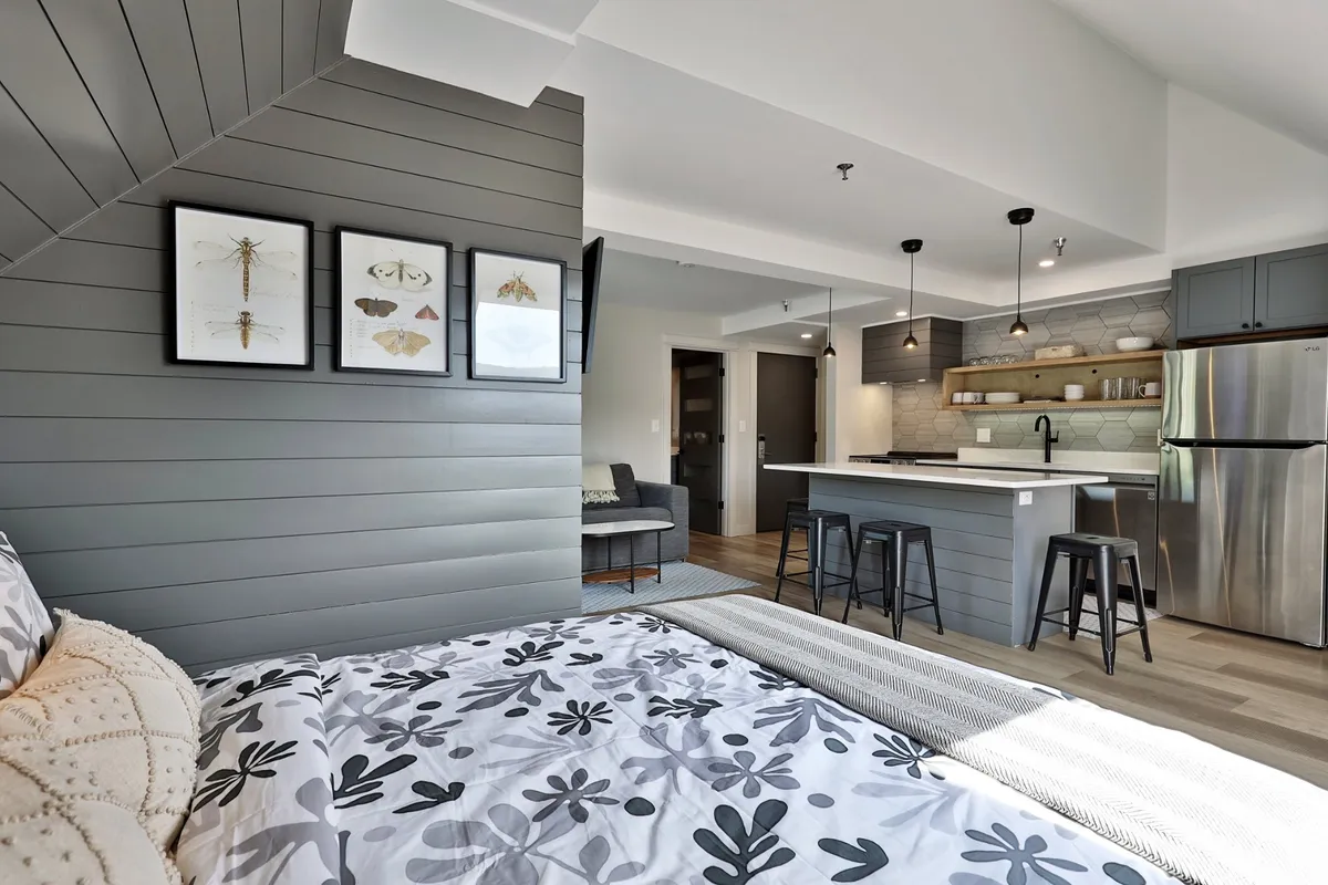 This image showcases a modern kitchen area complete with bar stools at the counter, blending into a cozy living space with a bed featuring floral bedding and framed artwork on the wall. The kitchen is well-equipped and inviting for meal preparation, while the living area offers a comfortable spot for relaxation or social activities.